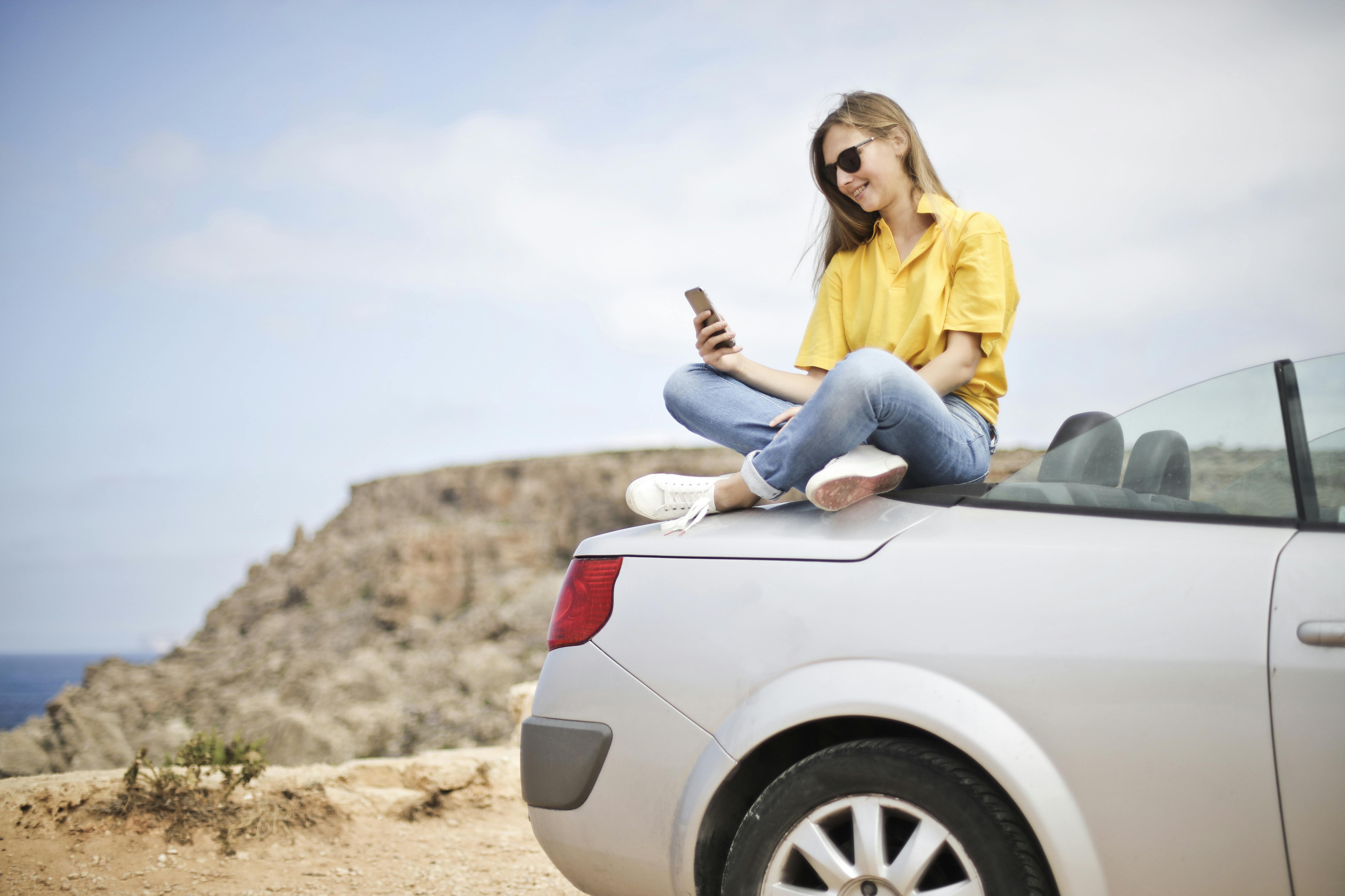 A girl wearing a yellow button-up shirt and jeans sits on the back of her car, looking at her phone in a remote desert location.